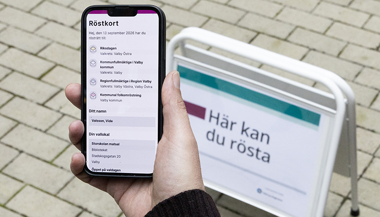 Hand med mobil framför skylt med texten här kan du rösta.